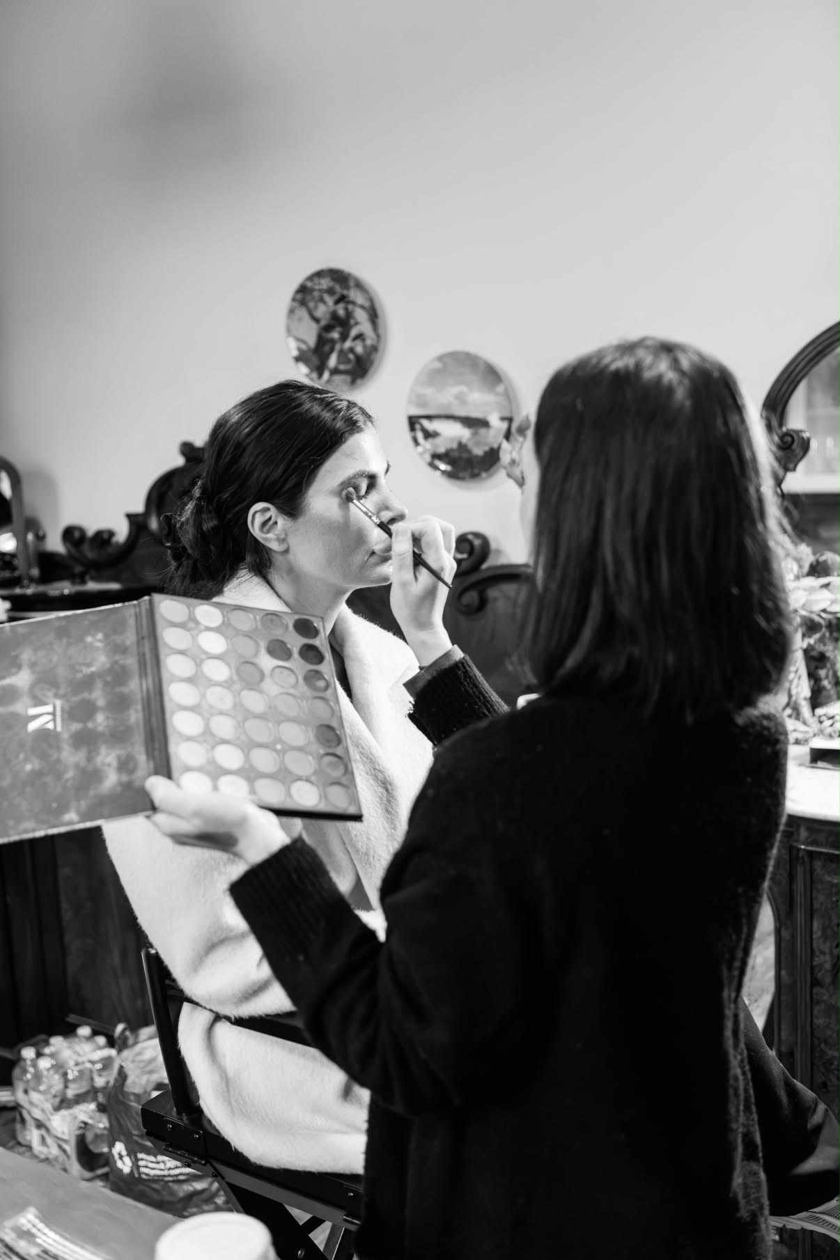 Black and white behind-the-scenes shot: J applying eyeshadow to a client, palette in hand