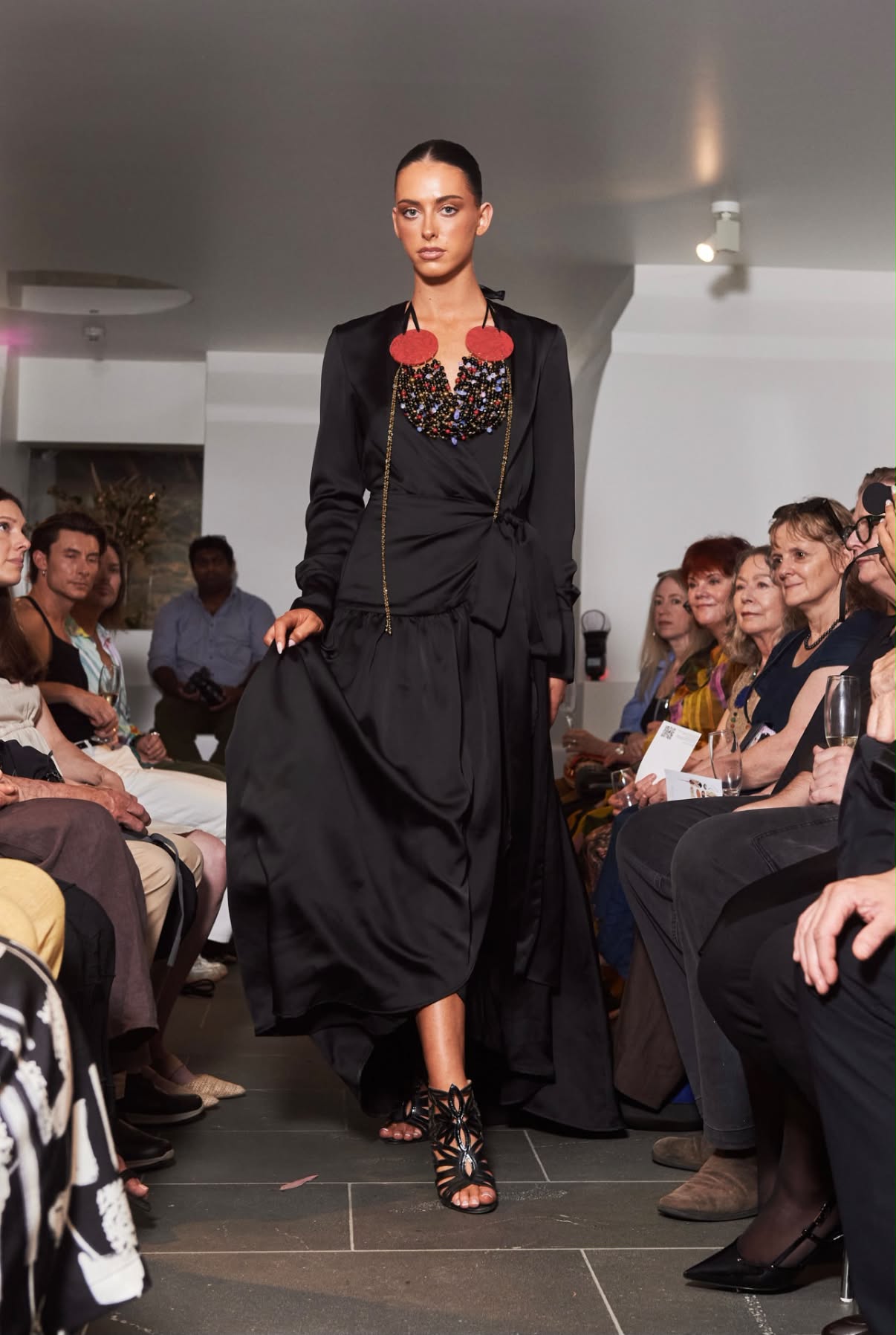 Runway: model walking in a black satin gown and statement necklace, bronzed skin and a sleek bun